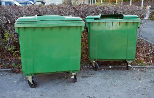 Two large green wheeled rubbish bins made of durable plastic are positioned side by side on a paved surface, with one being slightly larger than the other. The bins feature hinged lids on top, which are closed, and each has four small wheels for mobility. They are placed outdoors, possibly on a driveway or near a bin storage area, with a background of shrubbery and a metal fence, indicating a residential or commercial setting in Queens Park. The surface under the bins appears clean, and the bins show minimal signs of wear or weathering, reflecting their use for waste collection or rubbish disposal services associated with Office Clearance Queens Park. The natural outdoor lighting highlights the smooth texture and uniform green colour of the bins, emphasizing their functionality in rubbish removal activities in the local area.