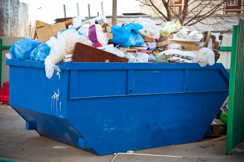 A large blue open-top skip filled to capacity with mixed household and office waste, including cardboard boxes, crumpled paper, plastic bags, and various discarded materials. The skip is situated outdoors on a paved surface, with a green metal fence to the right and a background of blurred urban buildings and trees. The waste materials are piled haphazardly, with some items spilling over the edges, and the textures of the materials include the smooth surfaces of cardboard and plastic, alongside the crinkled paper and soft plastic bags. The scene is illuminated by natural daylight, suggesting a clear day. This image relates to rubbish removal services provided by Office Clearance Queens Park for residential or commercial property clearances in the Queen's Park area, close to postal code NW10, highlighting the typical waste collection and disposal tasks undertaken in the local community.