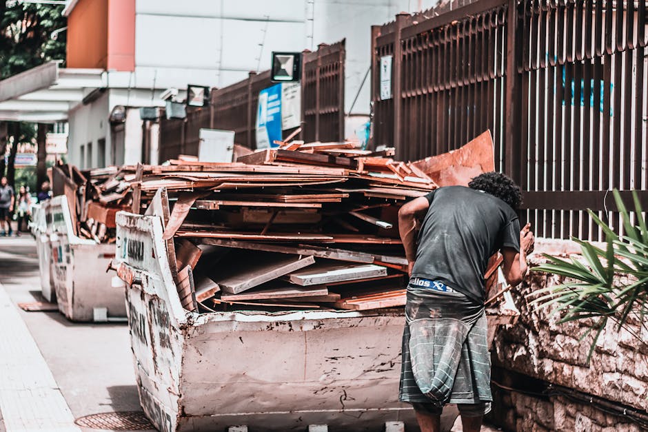 A vintage, brown upholstered sofa with wooden carvings on the armrests and backrest is positioned against a large, densely packed wall of assorted waste materials, including crumpled and flattened car