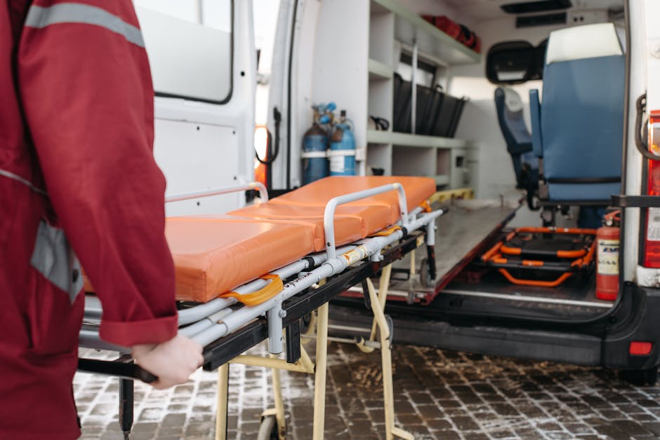 Inside the rear of an emergency medical response van, a person in a red uniform is seen loading a stretcher with an orange foam mattress onto a wheeled trolley. The stretcher features a metal frame an