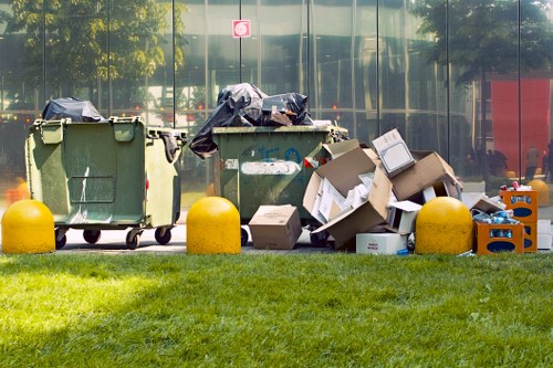 Two large green wheeled refuse bins with black plastic bags covering the tops are positioned on a paved surface, alongside several open cardboard boxes and miscellaneous waste materials. The boxes include a mix of brown and white cardboard, some flattened and others partially collapsed, with various items spilled around them. The waste appears to consist of packaging, plastic bottles, and other discarded items. Bright yellow plastic bollards are situated in front of the bins and boxes, separating the waste area from a grassy verge in the foreground. Behind the bins, a reflective glass building façade surfaces, with faint reflections of trees and urban surroundings visible. The scene is illuminated by natural daylight, creating a clear, professional snapshot of a typical outdoor rubbish collection or clearance setup often handled by companies such as Office Clearance Queens Park in the local area. The environment suggests an urban office or commercial area, with the waste awaiting removal or disposal, aligning with ongoing rubbish clearance services in the Queens Park district.