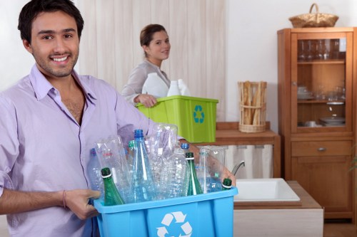A young man with dark hair and a trimmed beard, wearing a light purple shirt, is smiling and holding a large blue recycling bin filled with empty plastic bottles, including clear and green glass bottles, in a kitchen setting. In the background, a woman with dark hair tied back, wearing a light grey top, stands near a green rubbish recycling bin with a blue recycling symbol, placed on a wooden countertop. The kitchen features a white tile backsplash, a white sink, and wooden cabinets, with one cabinet partially open revealing glassware inside. The setting is well-lit by natural light, and the scene reflects a domestic environment focused on sorting waste materials for recycling, highlighting efficient rubbish management and eco-friendly practices consistent with professional waste disposal services in Queens Park.