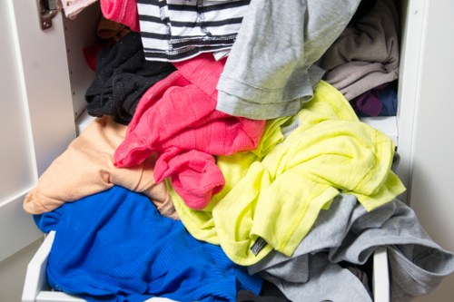 A drawer filled with a disorganized pile of mixed clothing items, including brightly coloured and neutral-toned garments such as pink, yellow, blue, grey, and beige, with various textures and fabrics visible. The clothes are piled haphazardly, spilling out of the drawer onto the surrounding surface, and the background shows a glimpse of a white or light-colored interior, suggesting a domestic setting. This image illustrates typical clothing mess often encountered during household rubbish removal or clearance services, which companies like Office Clearance Queens Park may help manage in the Queens Park area, postcode inferred from the page context. The cluttered state of the drawer emphasizes the need for effective rubbish clearance and organisation, matching the service category focus.